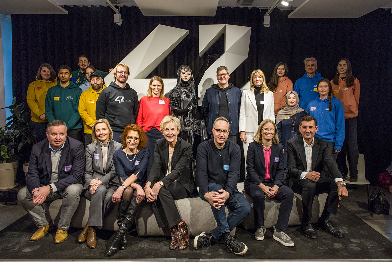 Das Foto zeigt eine Gruppe an Menschen stehend und sitzend vor einer dunklen Wand mit der Aufschrift 42.