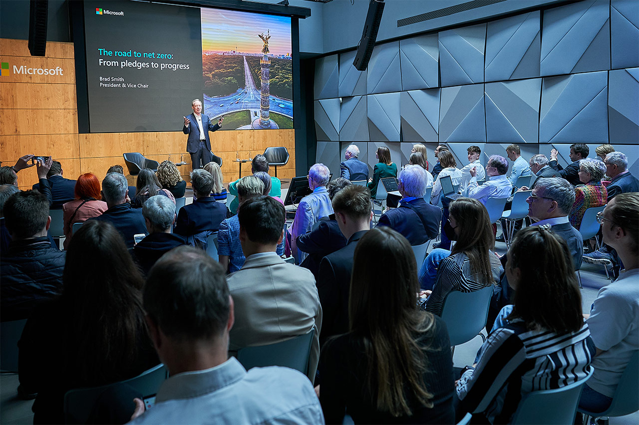 Das Foto zeigt Brad Smith auf der Bühne des Atriums von Microsoft Berlin während er seinen Vortrag hält. Im Publikum sitzen viele Menschen, die ihm gespannt zuhören.
