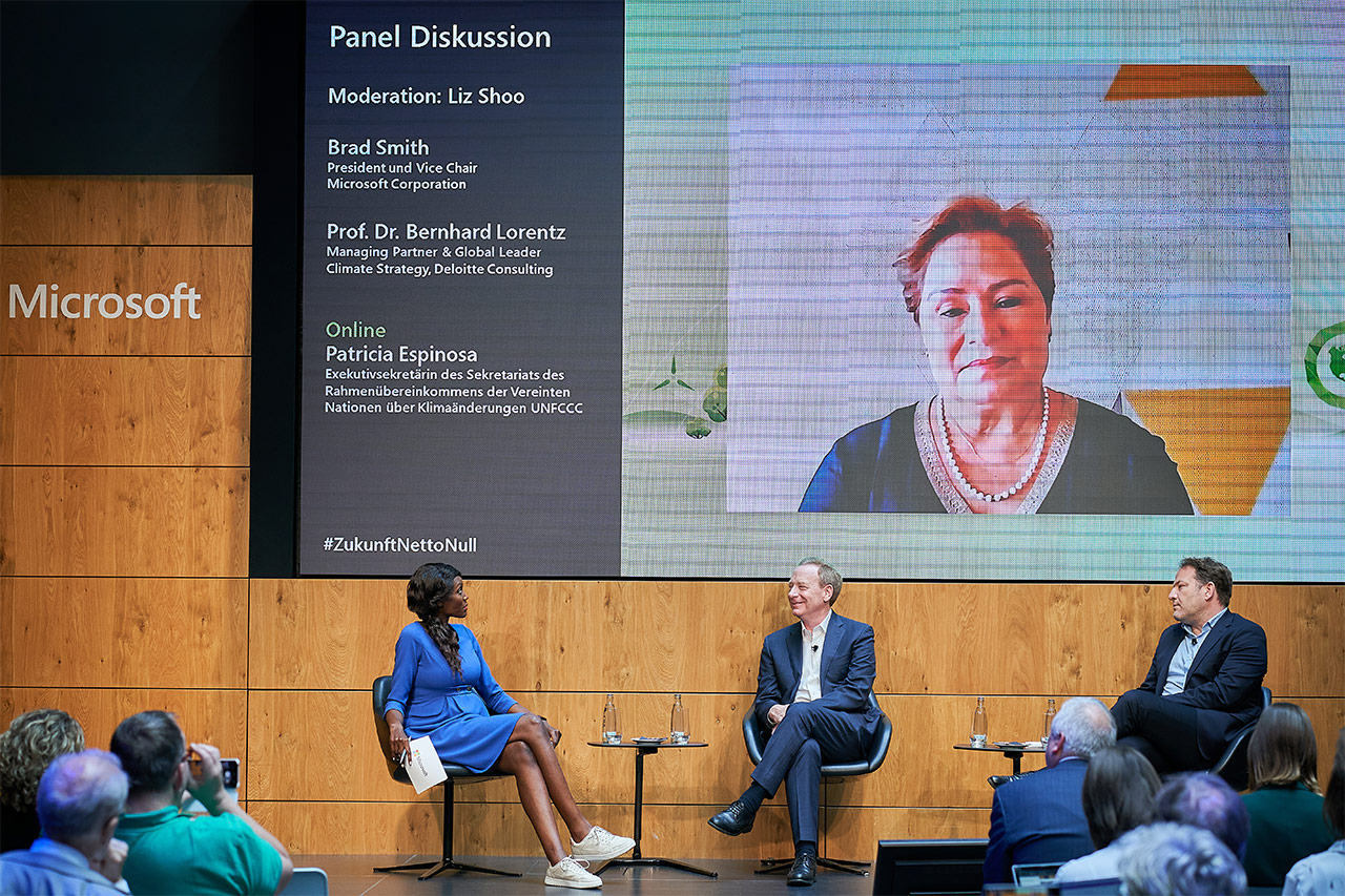 Das Foto zeigt die Moderatorin Liz Shoo, Brad Smith und Prof. Dr. Bernhard Lorentz auf der Bühne vom Atrium von Microsoft Berlin sitzend. Im Hintergrund sieht man auf dem Bildschirm Patricia Espinosa, die der Paneldiskussion zugeschaltet war.