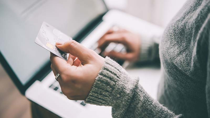 Eine Person bedient mit ihrer rechten Hand einen Laptop, während ihre linke eine Bankkarte hält.