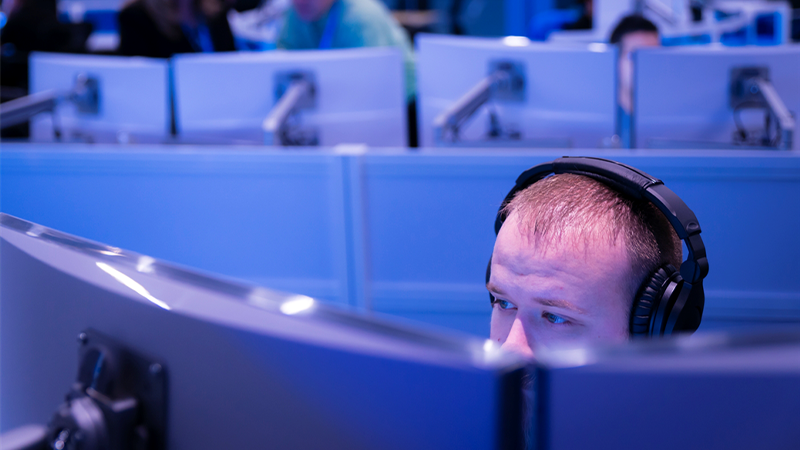 Das Bild zeigt einen Mitarbeiter mit Kopfh&ouml;rern auf einen Bildschirm blickend. Im Hintergrund befinden sich weitere Mitarbeiter vor Computern