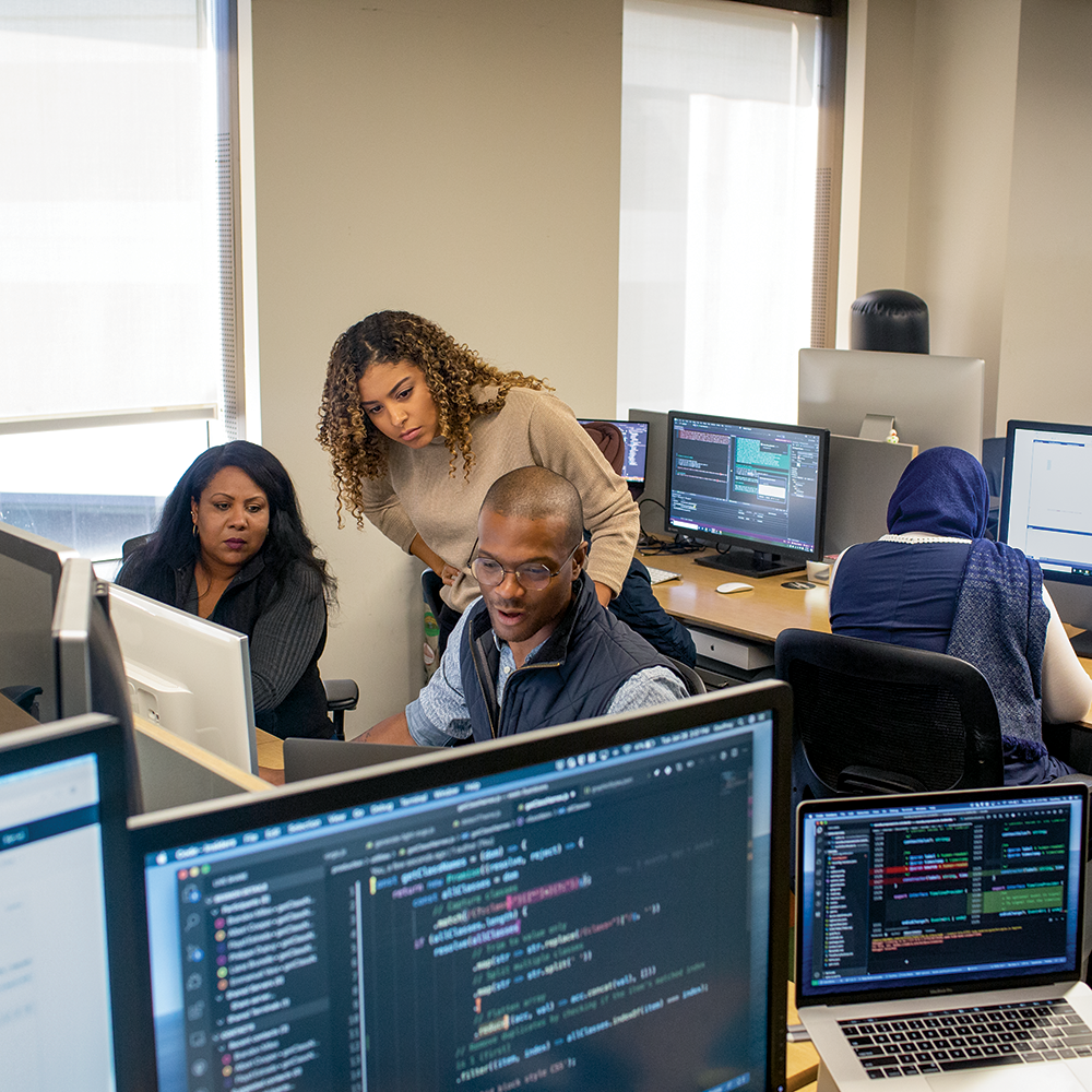 Three people gathered around a computer in an office space filled with multiple monitors.