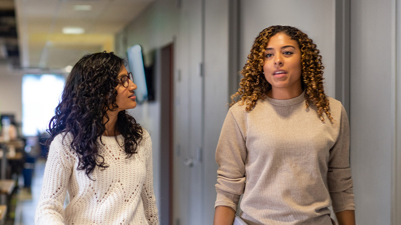 Two women walking together in an office hallway, talking as they walk.