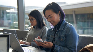 Two people sitting indoors by large windows, working on laptops, with one person also using a smartphone and wearing earbuds.