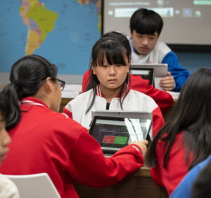 The camera focuses on a girl student in a classroom full of students.