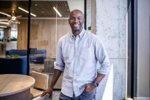 Black man standing and smiling in a office in a blue collared shirt.