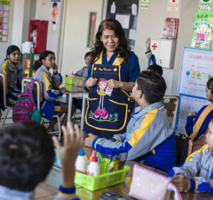 A female teacher stands amidst students in a classroom.