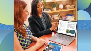 Two education leaders sitting at a table in a school office and looking at the screen of a laptop together.