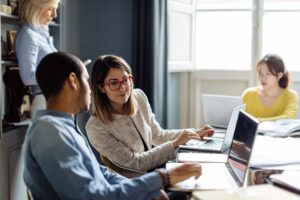 Small group of professionals collaborating at a table with laptops and notebooks, discussing ideas in a bright office meeting space.