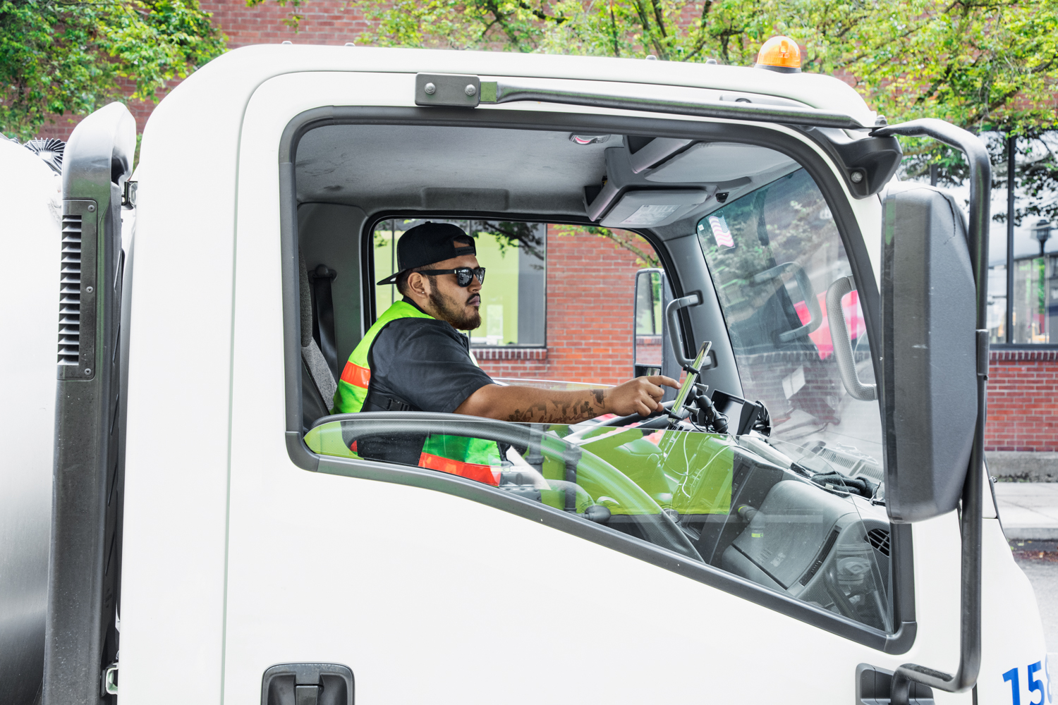 Male worker sitting in driver&lsquo;s seat of small loading truck using tablet mounted on dashboard. Photo taken from outside passenger door. Man&lsquo;s tattoos visible on arm.