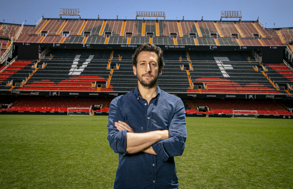 A portrait of Franco Segarra in a blue shirt standing in an empty soccer stadium.
