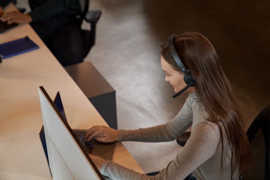 A woman sitting at a desk, using a laptop, wearing a headset.