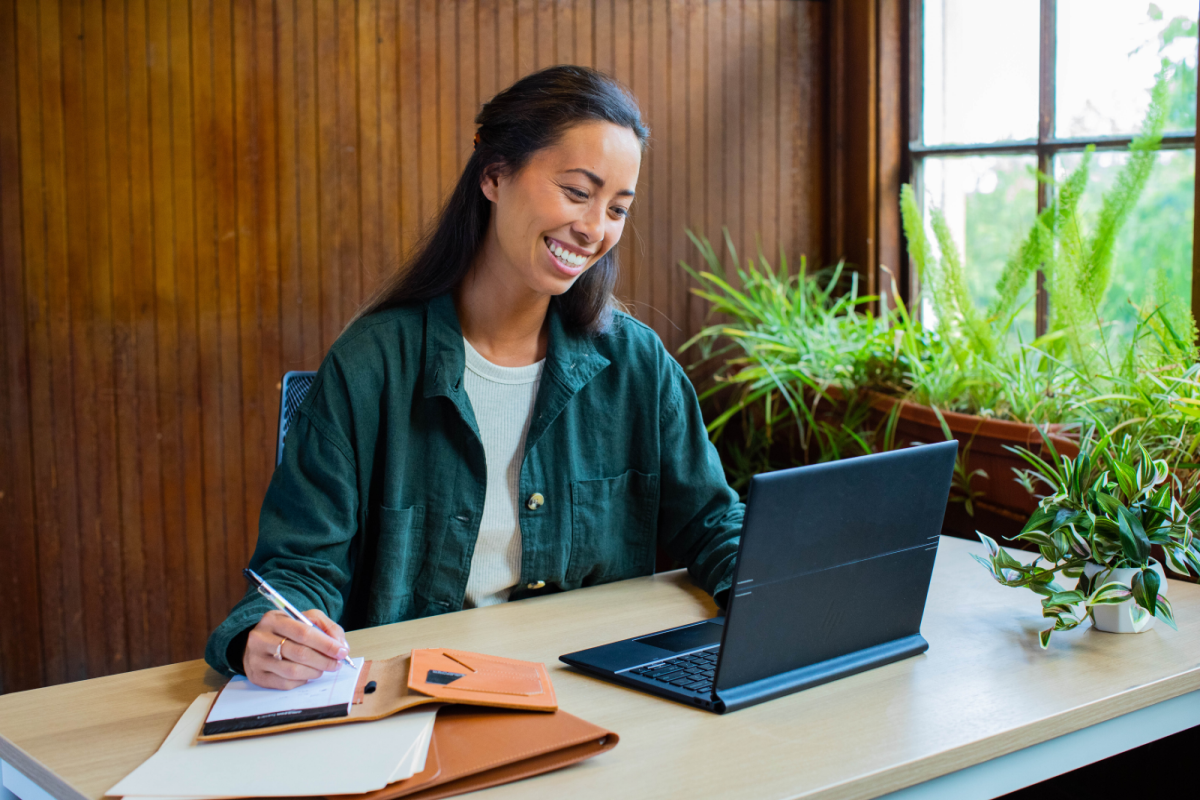Hybrid work. Woman sitting at casual desk at home with an HP Elite Folio which is a commercial device. Taking notes and smiling. Windows 11 Pro.