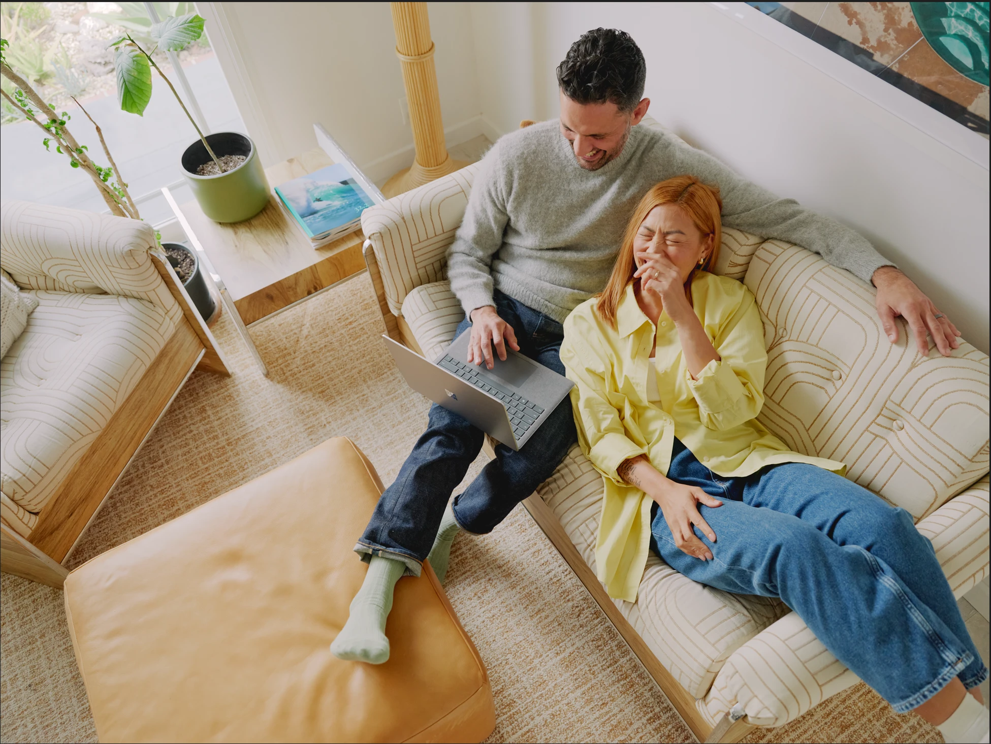Microsoft 365 lifestyle photography of a male and female adult on a couch smiling, laughing and looking at a Surface Laptop 5.