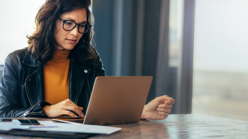 Asian business woman sitting at a table in an office, typing on her laptop with focus. Young female professional showing a dedication and commitment to her project at work.
