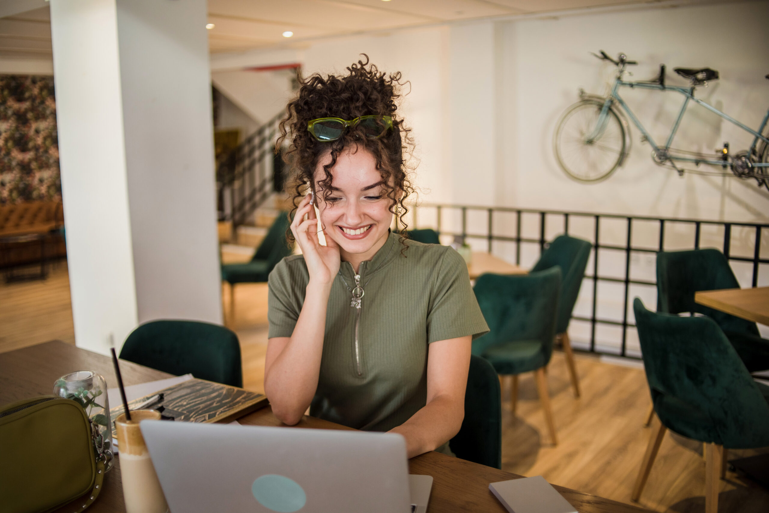 A young woman is working on a laptop while sitting in a cafe and drinking a frappe coffee that is on the table.