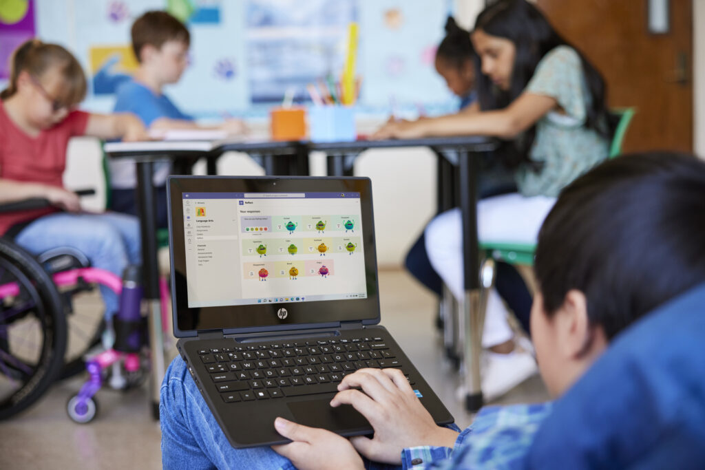 A student sitting on a beanbag in a classroom working on a laptop while a group of fellow students work at a table in the background.