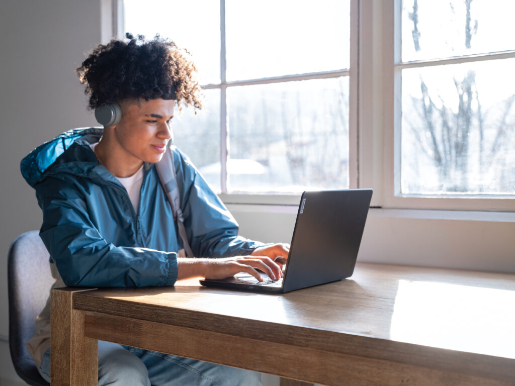 High school aged student wearing headphones and working on a laptop at a table near a window.