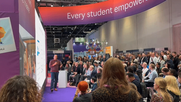 GIF. A conference presenter speaks to an audience of approximately 75-100 people in a crowded conference hall.