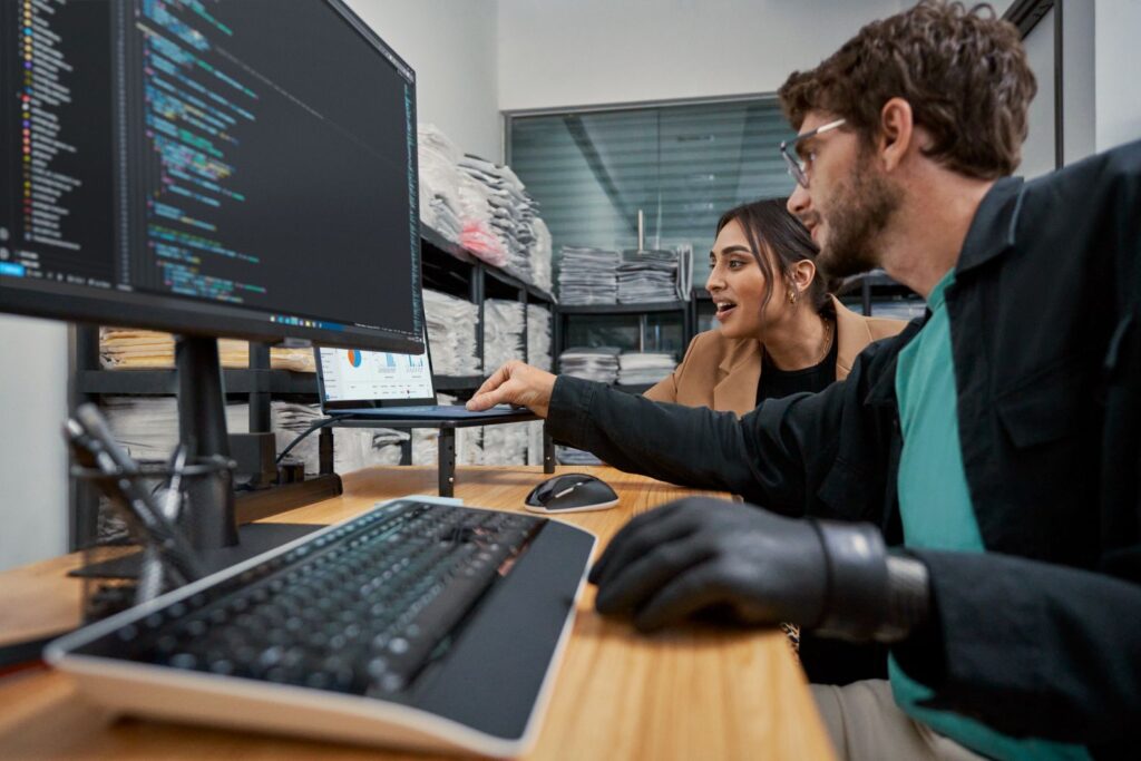 Two people collaborate at a workstation with a laptop and external monitor.
