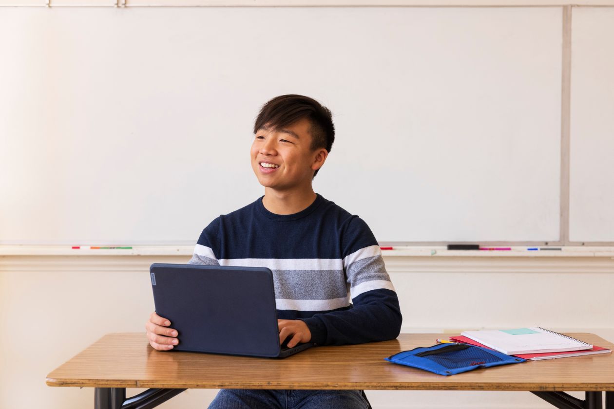 An older student sitting at a table in a school classroom while using a laptop.