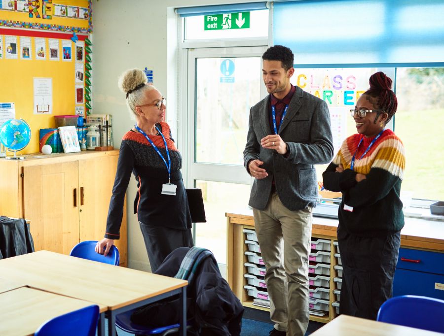 Three educators stand together and talk with each other in a school classroom.