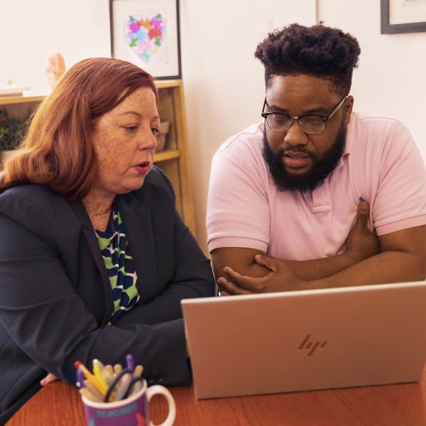Two educators sitting at a table and working together on a laptop in a school office.