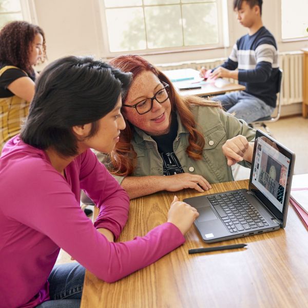 An educator in a classroom helps a student with schoolwork on her laptop.