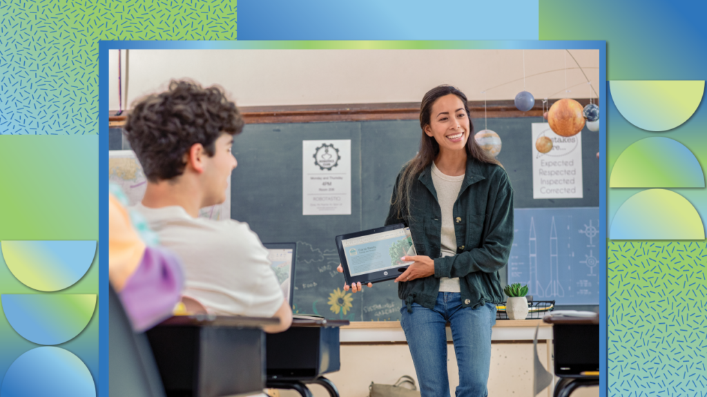 A teacher holding a folded laptop in front of students in a classroom.
