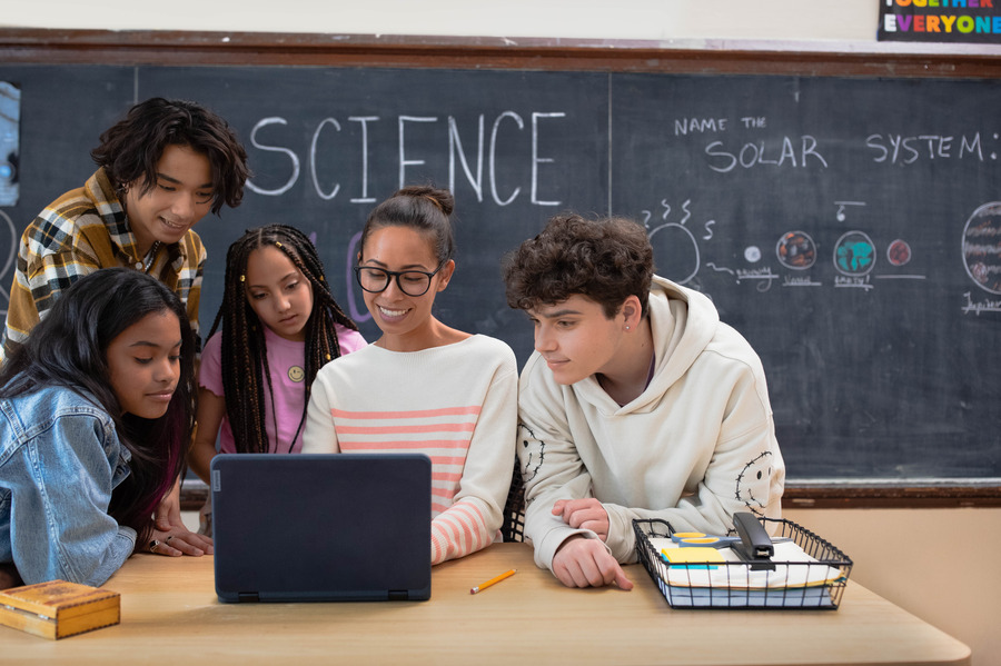 An educator and four students looking at a laptop at the educator’s desk in a school classroom.