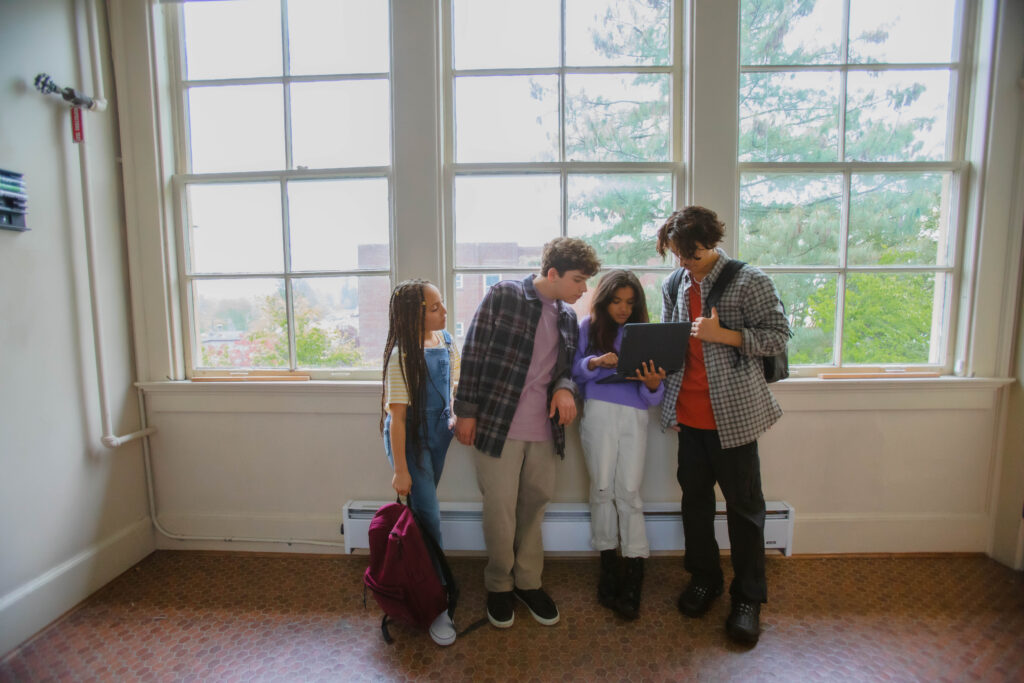 A group of four middle school students stand in a school hallway looking at an open laptop screen. 