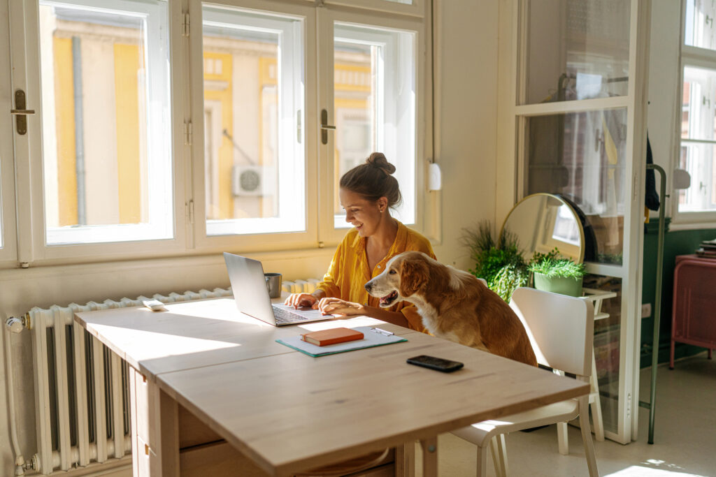 A woman sitting at a table at home and working on a laptop. A dog sits next to her.