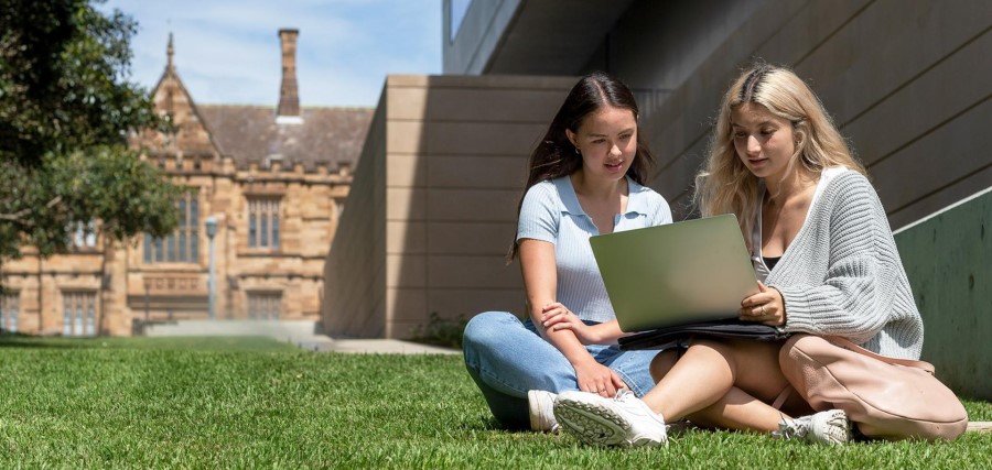Two female college students working on laptop together while sitting in the grass in a university garden.
