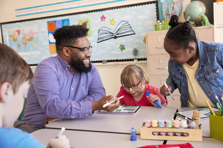 A teacher sitting at a table and engaging with younger students who are working on laptops.