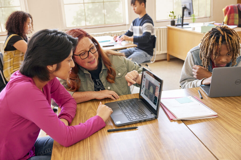 A teacher working on a laptop with students in a classroom setting.