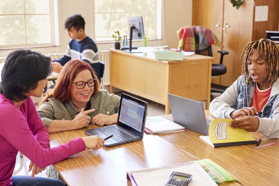 A classroom scene with students at communal desks and an educator leans in to help one of the students.