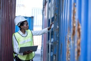 Female employee in hard hat and safety vest inspects shipping container.