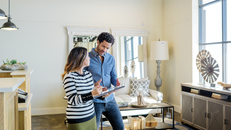 Eine Mitarbeiterin eines Einzelhandelsgeschäfts, berät einen Kunden. Als Hilfsmittel nutzt sie ein Tablet. Der Kunde schaut interessiert auf das Gerät.