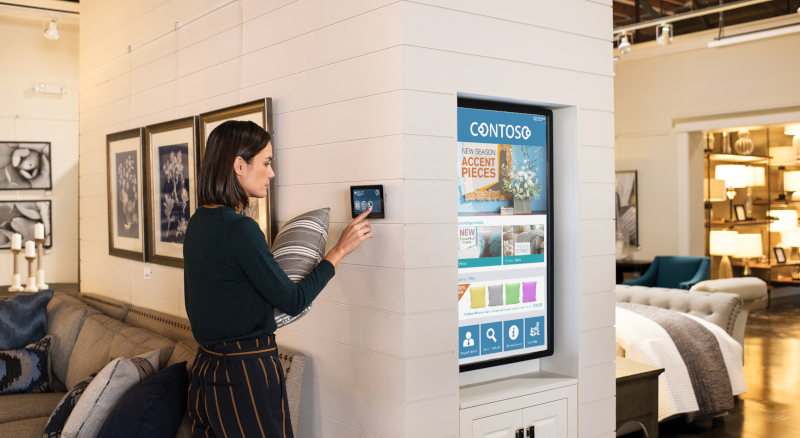 Eine Kundin steht in einem Einzelhandelsgeschäft, hält ein Kissen und benutzt ein an der Wand angebrachtes Tablet.