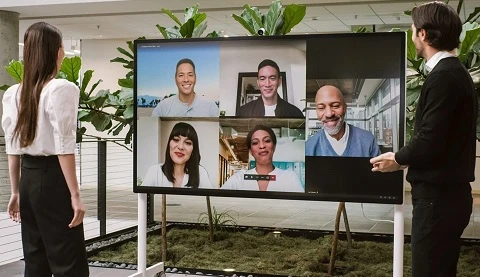 Zwei Personen stehen vor einem gro&szlig;en Bildschrim auf dem andere Personen zu sehen sind. 