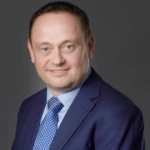 Photo of Phil Hadfield, a man with dark hair in a navy business suit with a light blue shirt and blue tie smiling at the camera