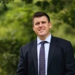 James Featherstone, a man wearing a suit and tie smiling at the camera. He is outside in front of some green bushes.