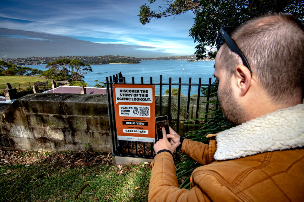 A photo of a man using their phone to scan a Hello Lamp Post photo, which says it can give them the story of the scenic lookout they are at.