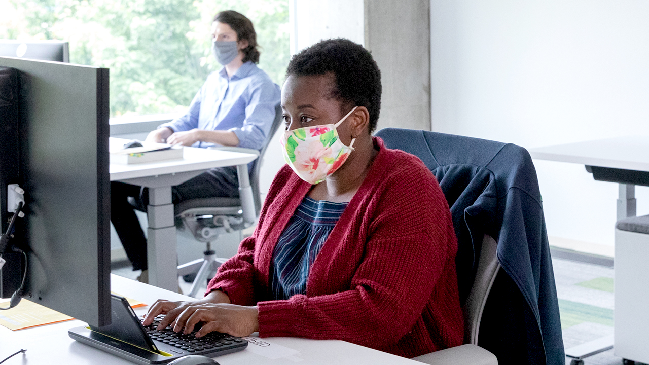 Woman wearing a mask, sitting at a desk and using a keyboard to type.