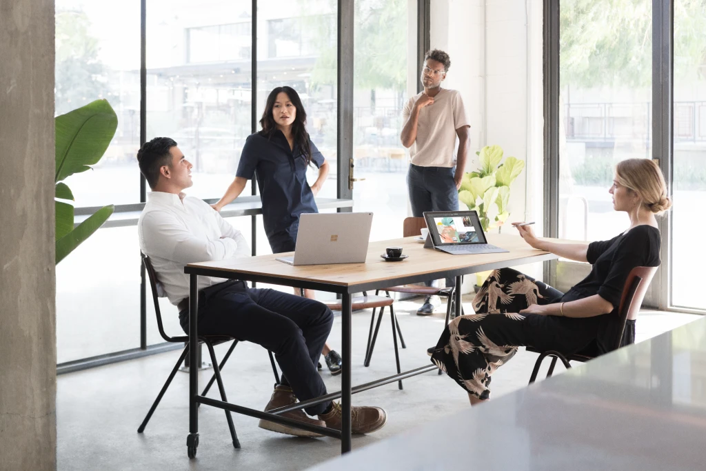 A group of people gathered in a room at work with their computers is talking to each other