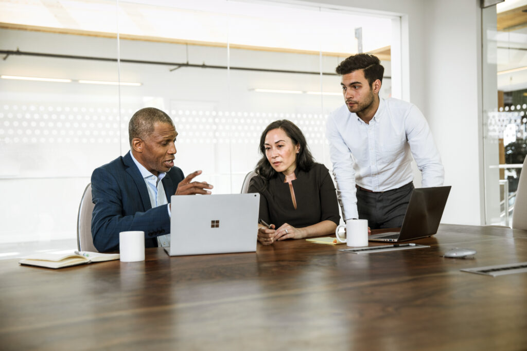 Two male and one female corporate office workers sitting at conference room table in discussion. There are two open laptops on the table.