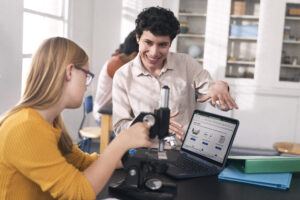 Female K-12 student and female educator in science classroom next to small microscope looking Lenovo 14w gen 2. Male student works at separate table in the background.
