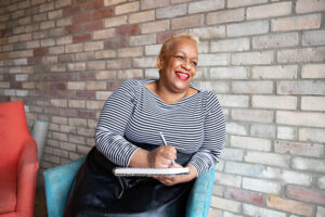 An mature black entrepreneur holding a notebook sits in an armchair against a brick wall in a cafe.