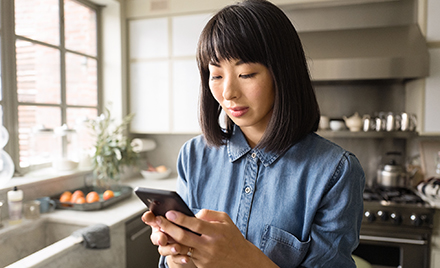 A woman uses a phone next to a window in her kitchen.