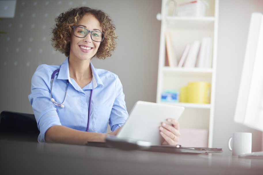 Woman wearing a stethoscope, sitting at a desk, and holding a tablet